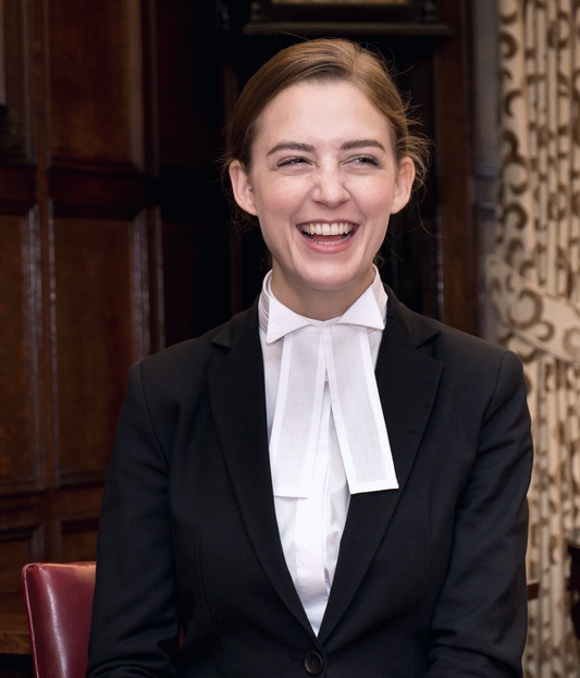A young female barrister stands in an office setting, confidently smiling. She wears a dark suit complete with a tunic shirt, wing collar and barrister bands.